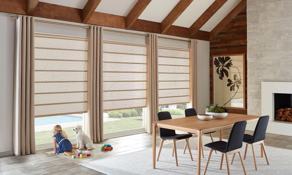 Cozy living room with large windows covered by beige Roman shades, featuring a wooden dining table, black chairs, and a child playing with a dog near the window.