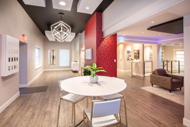 A modern showroom with a bright geometric chandelier, red accent wall, and light-colored floors, featuring a sleek round table and chairs in the foreground.