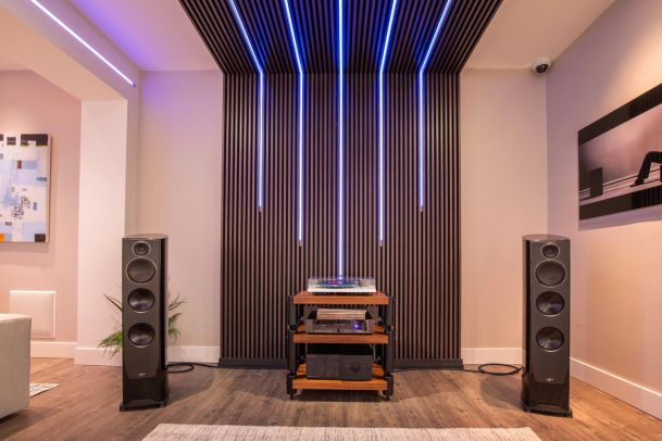 A home audio setup featuring large tower speakers flanking a wooden audio rack with electronics, set against a modern wall with vertical wooden slats and integrated blue LED lighting.